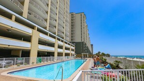 Une piscine située à côté d’un immeuble de grande hauteur, doté de nombreux balcons, offrant une vue sur la plage et comprenant un espace clos avec des chaises de plage.