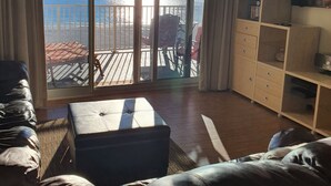 Un salon doté d’une grande porte-fenêtre coulissante donnant sur un balcon avec vue sur la mer. On y trouve un ventilateur de plafond, un canapé, une table basse et un meuble pour la télévision, sur lequel est posé un téléviseur à écran plat.