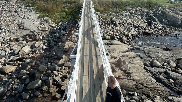 Un puente de madera que lleva a una casa blanca con porche, rodeada por una costa rocosa y un bosque.