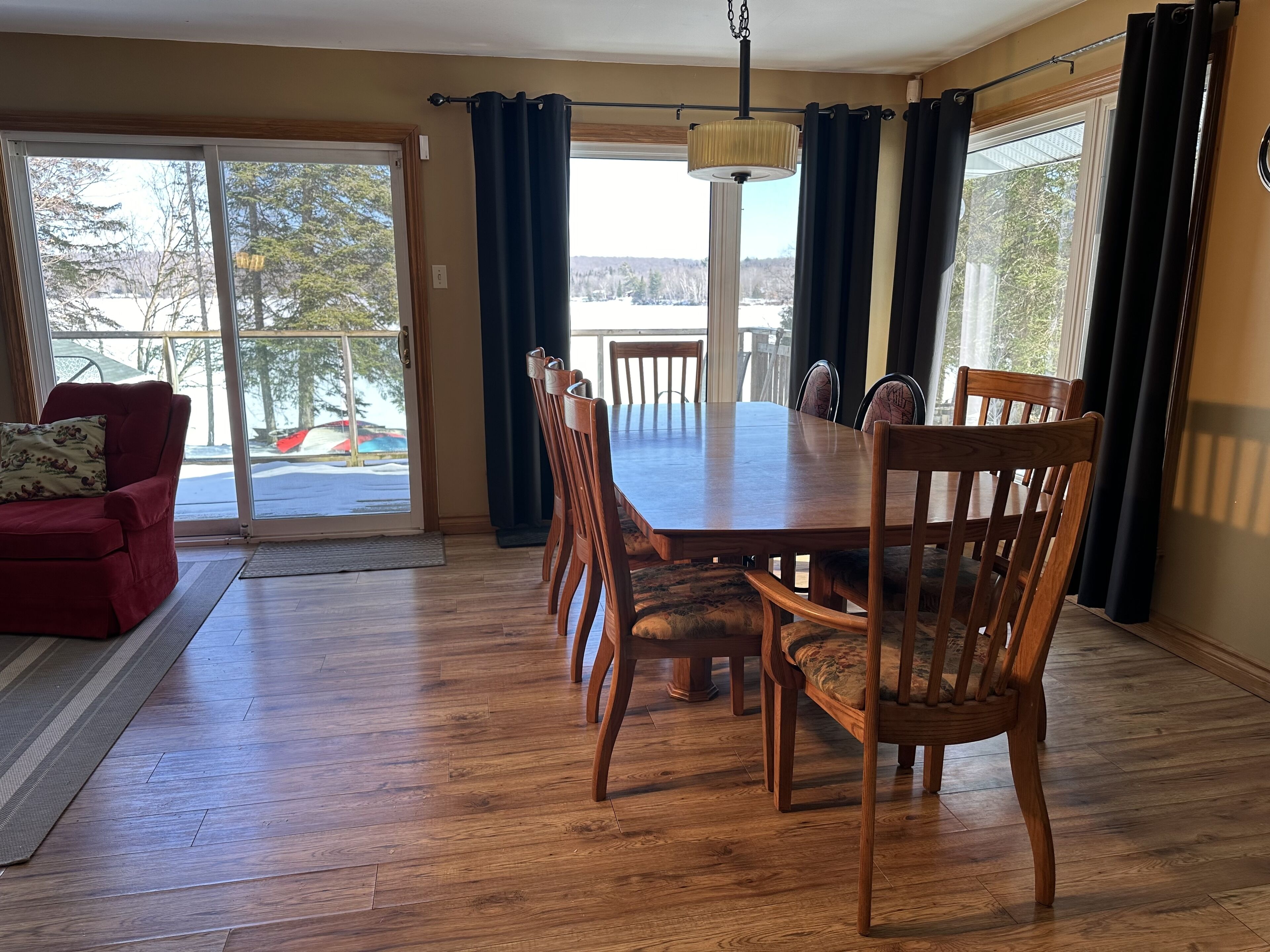 A dining area with a wooden table and chairs, a red armchair, and a view through large windows.