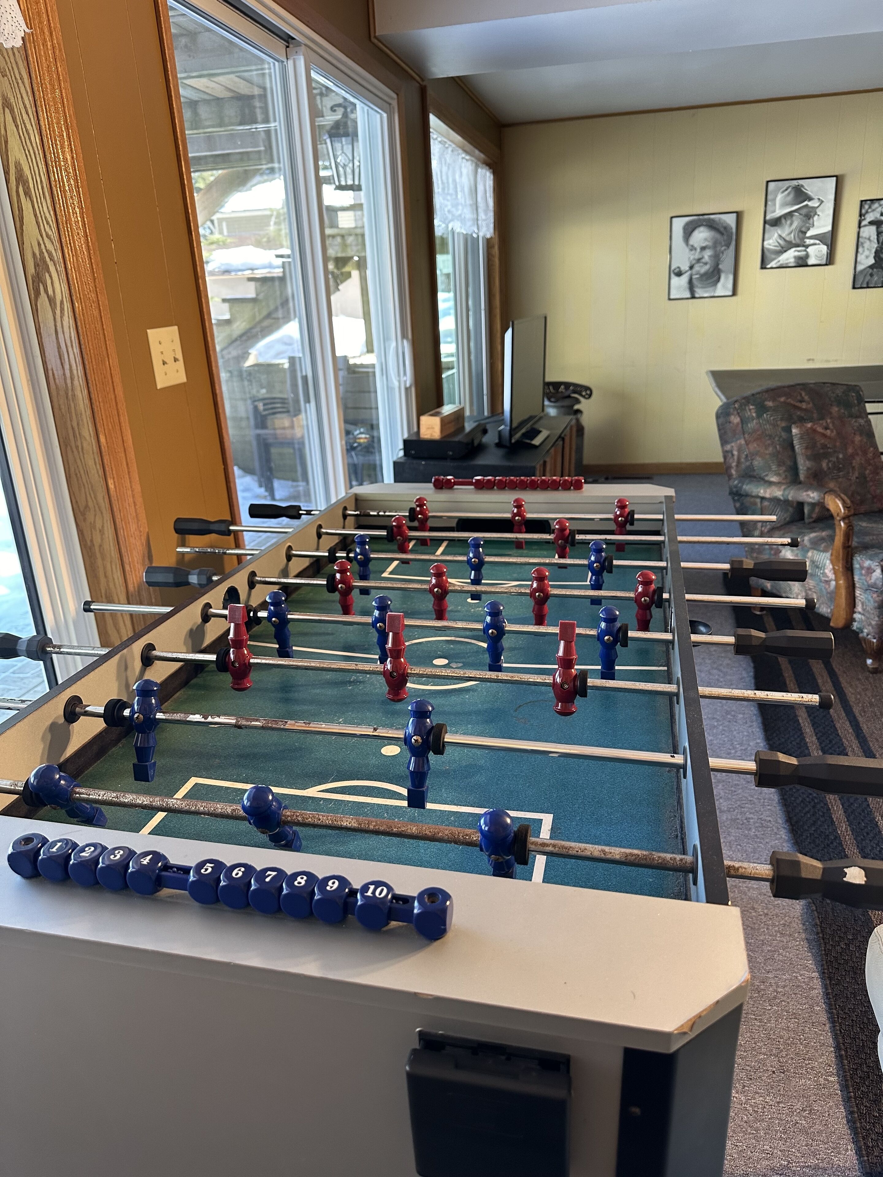 A foosball table in a room with a large window, a television, and framed pictures on the wall.