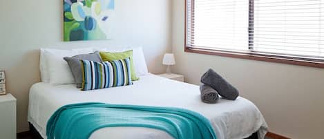 Uma cama bem arrumada com um manta verde-água, almofadas listradas e uma toalha enrolada ao lado.