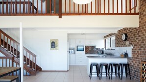 A spacious kitchen with a central island, a dining area with stools, and a staircase leading to an upper level. - A stone's throw from Mollymook Beach (Mollymook)