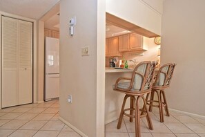 A kitchen with a refrigerator, wooden cabinets, a clock, and two wicker chairs. - Doveplum 721- 2 bedroom 2.5 bath overlooking serene canal with private beach access (Siesta Key)