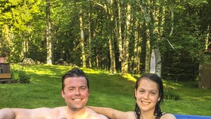 Um casal em uma banheira de hidromassagem, com gramado e árvores ao fundo.