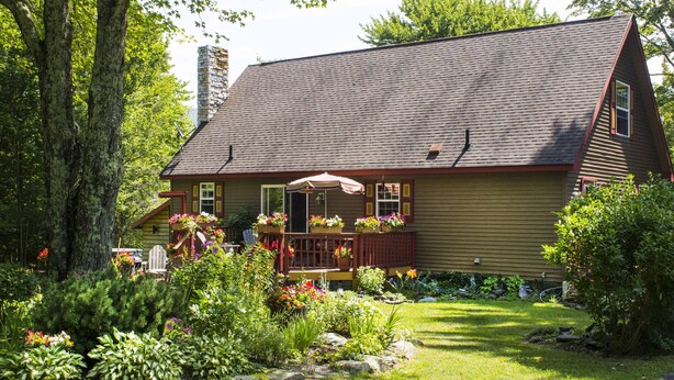Uma casa com telhado inclinado, varanda frontal com guarda-corpo vermelho e jardim com diversas plantas e flores.
