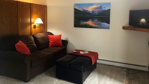 Uma sala de estar com um sofá marrom, almofadas vermelhas, um banquinho preto, uma pintura de uma paisagem montanhosa e uma TV sobre uma estante de madeira.