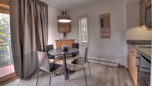 Uma área de refeições com mesa redonda, cadeiras, luminária pendente e vista para o exterior através de uma janela com persianas.