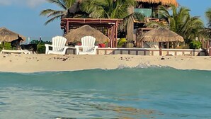 Una propiedad frente a la playa con un edificio de techo de paja, sillas de descanso blancas y palmeras.