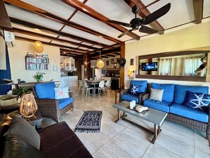A spacious living area with a sofa, coffee table, and a dining table. There is a ceiling fan, a kitchen in the background, and a television mounted on the wall.
