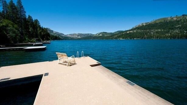 A wooden dock with lounge chairs and a pool, overlooking a calm lake with boats and mountains in the background.
