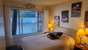 A hotel room with a bed, two bedside lamps, a window with a view of trees and water, and artwork on the wall.