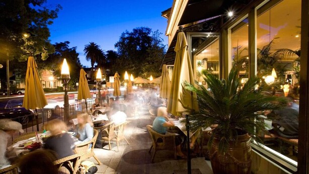An outdoor dining area with wicker chairs and yellow umbrellas, surrounded by palm trees and a building with large windows. - Vintage Flat | Mins to Golden 1 Center, Capitol (Sacramento)