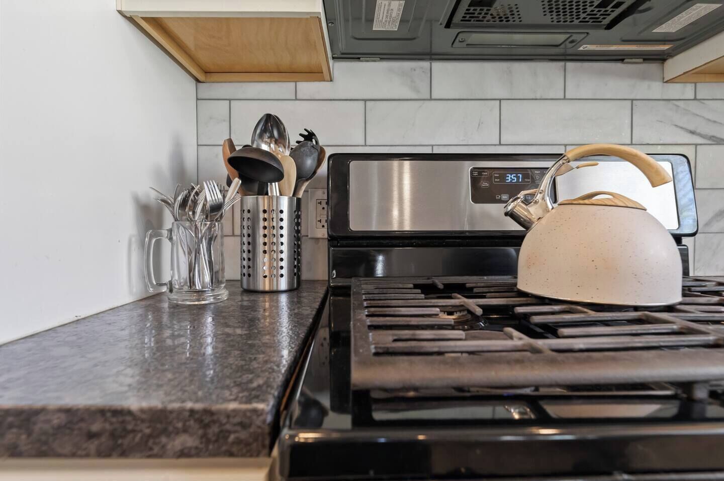 A kitchen with a stainless steel stove, a kettle, and utensils on the counter.