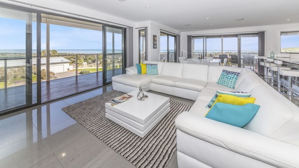 Un salon moderne avec un canapé d’angle blanc, une table basse et de grandes fenêtres du sol au plafond offrant une vue sur la mer et les bâtiments.