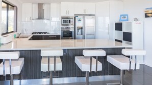 Une cuisine moderne avec des armoires blanches, un grand îlot central doté de tabourets, des appareils en acier inoxydable et une vue sur l’extérieur.