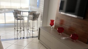 Un salon moderne avec un téléviseur à écran plat fixé à un mur en bois, une table à manger en verre avec des chaises en métal et un sol carrelé.