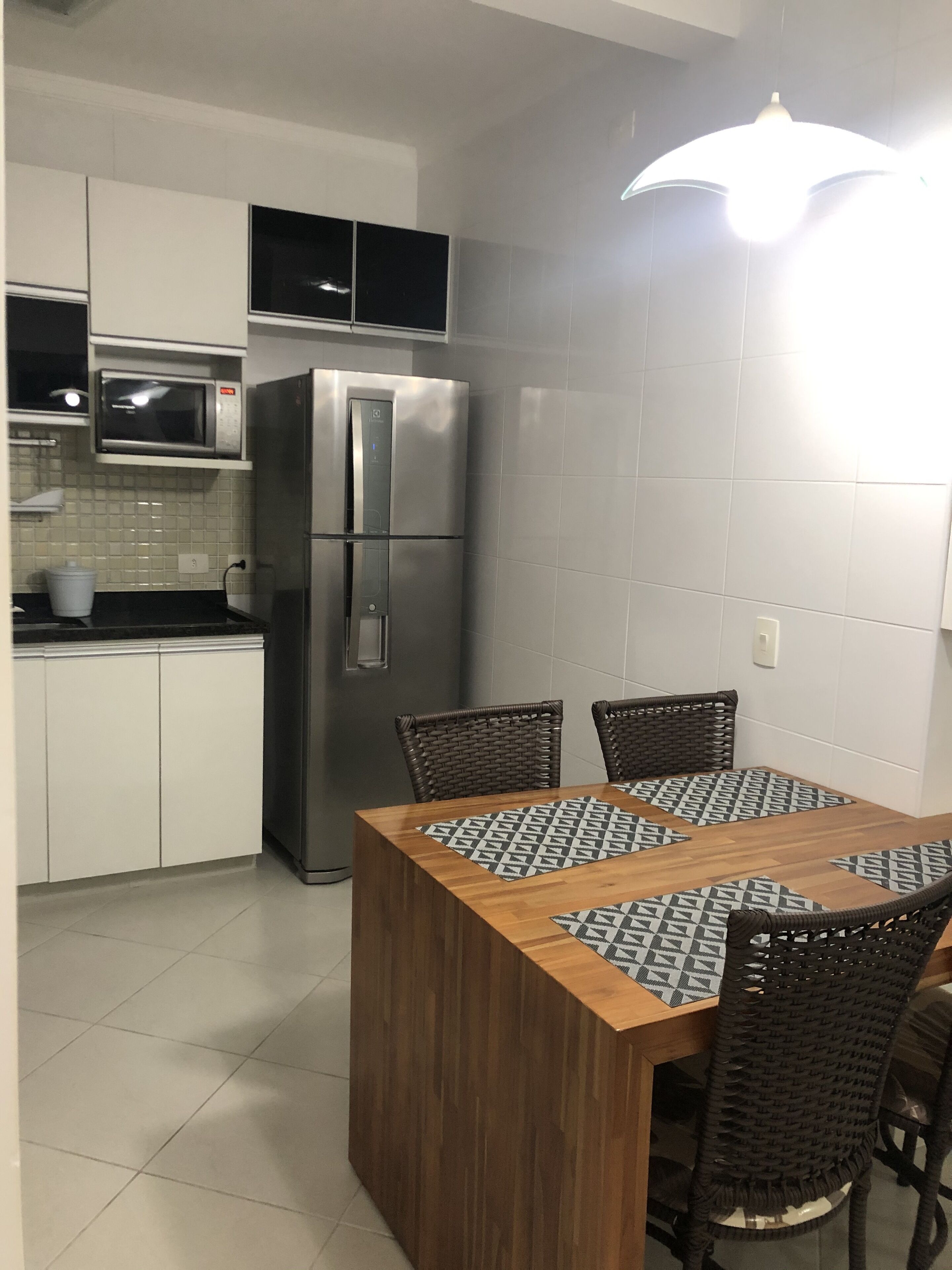 Een keuken met witte kastjes, een roestvrijstalen koelkast, een magnetron en een eettafel met geruit tafelonderleggers en rieten stoelen.