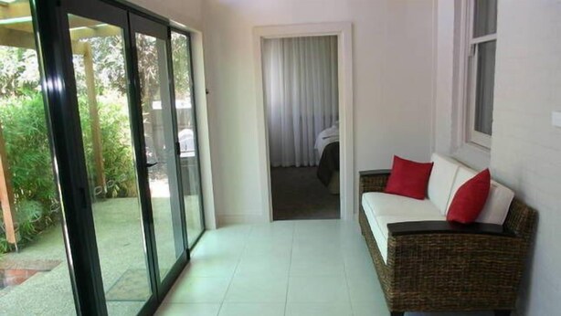 Una habitación con una puerta corredera de vidrio que da a un jardín, un sofá de mimbre con cojines rojos y una cama blanca al fondo.