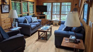 Interior acogedor de una cabaña con paredes de madera, un conjunto de sofás, un televisor y ventanas amplias que ofrecen vistas al bosque.