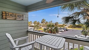Ein Holzschild mit der Aufschrift 'Beach House' an einer Balkonbrüstung, mit Blick auf den Strand und Palmen.