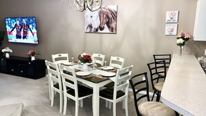 A dining area with a round table set for six, surrounded by white chairs. A chandelier hangs above, and a television is mounted on the wall.