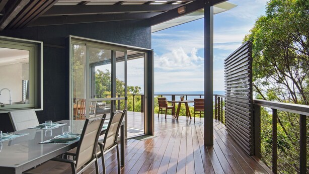 A modern outdoor dining area with a view of the ocean, wooden deck, and a glass-enclosed dining space.