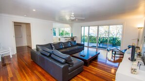 A modern living room with a leather sofa, wooden flooring, a coffee table, and a balcony with a view of greenery. - NICK'S PLACE - ON BEACH HOLIDAY HOUSE (AGNES WATER)