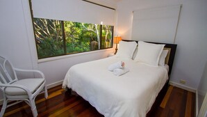 A neatly made bed with white linens and pillows, a white chair, and a window with a view of greenery. - NICK'S PLACE - ON BEACH HOLIDAY HOUSE (AGNES WATER)