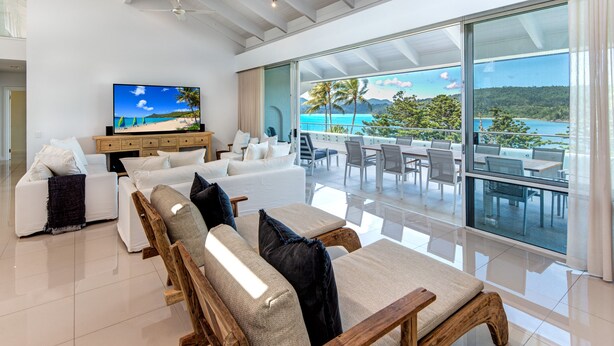 Una sala de estar moderna con un televisor grande, sofás blancos, muebles de madera y un balcón con vistas al mar.