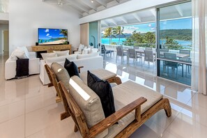 A modern living room with a large TV, white sofas, wooden furniture, and a balcony with a view of the sea.