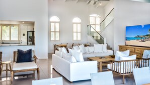 Amplia sala de estar con un sofá blanco, sillas de madera y una mesa de centro. Se observa una escalera que conduce a un nivel superior, y un televisor grande en la pared.