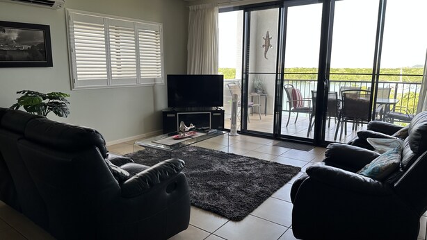 Un salon doté d’un téléviseur à écran plat, d’un canapé en cuir noir, d’une table basse en verre et offrant une vue sur un balcon aménagé avec du mobilier d’extérieur.