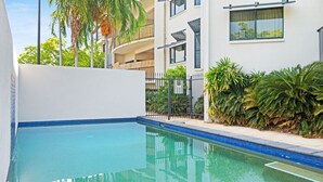 Une piscine dont le bord est recouvert de carreaux bleus, entourée d’un mur blanc et d’une clôture. On aperçoit des palmiers et d’autres plantes en arrière-plan.