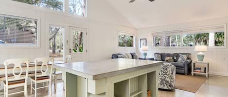 A spacious living area with a large central island, a ceiling fan, and a view of the outdoors through large windows.
