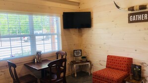 Un interior acogedor de cabaña con detalles de madera, un televisor montado en la pared, un letrero que dice 'GATHER', un sillón rojo con estampado y una mesa con sillas.