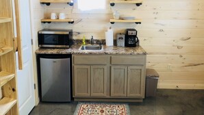 Una cocina compacta con microondas, cafetera y lavamanos. Hay armarios de madera y una pequeña alfombra en el suelo.