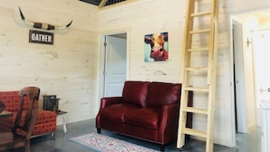 Una habitación acogedora con un sofá de cuero rojo, una escalera de madera, una obra de arte con la figura de una vaca y un letrero que dice 'Gather' en la pared.