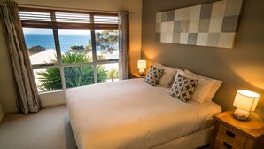 Ein Schlafzimmer mit einem großen Bett, zwei Nachttischen mit Lampen, Blick aufs Meer durch das Fenster und einem Wandbild mit Schachmuster.