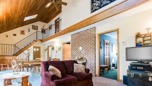 Un salón espacioso con techo alto, chimenea, escalera y un televisor sobre un aparador.