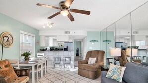 A spacious living room with a ceiling fan, a dining area with a table and chairs, a kitchen in the background, and a large mirror reflecting the room.