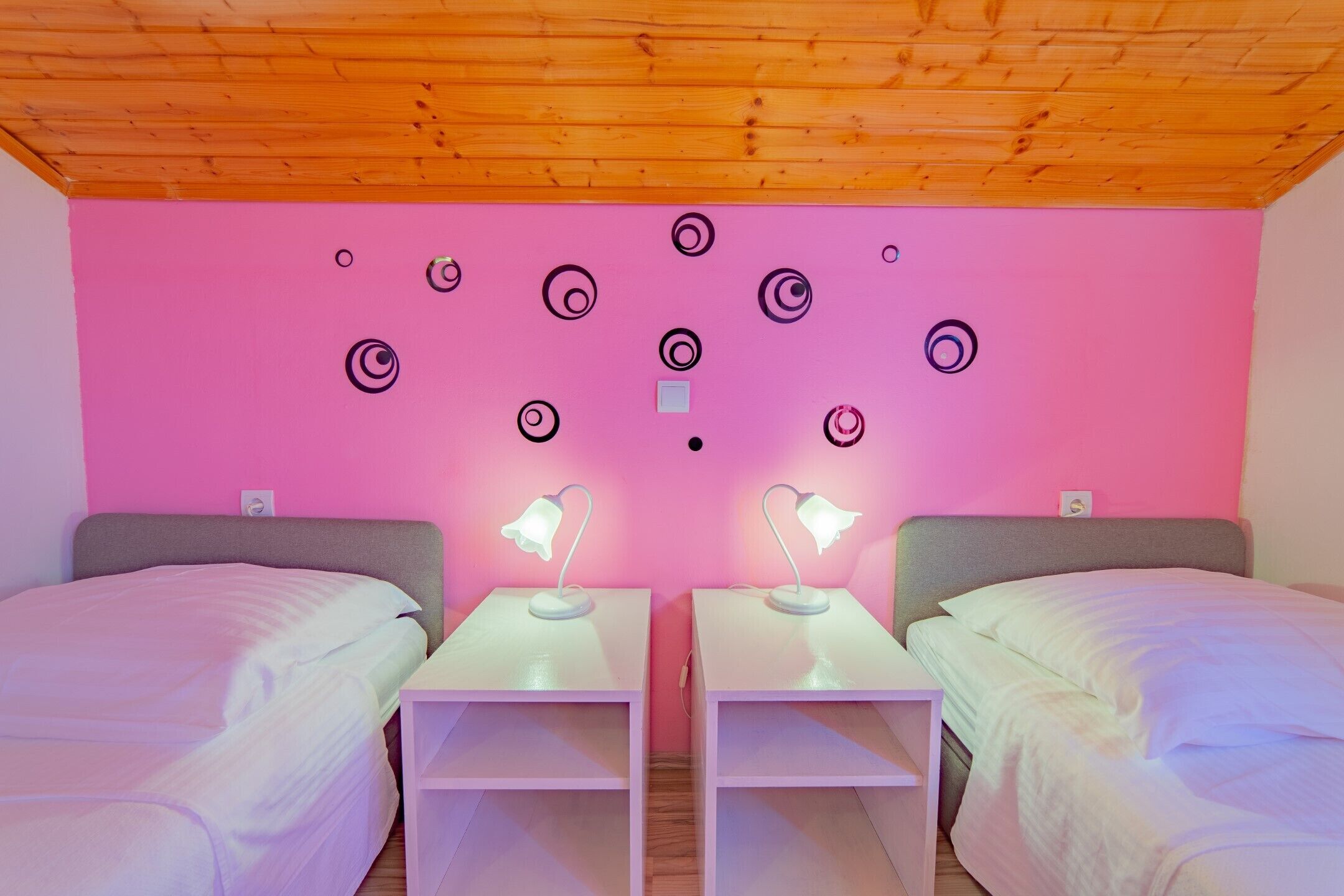 A room with two beds, a wooden ceiling, and a pink wall with circular patterns.