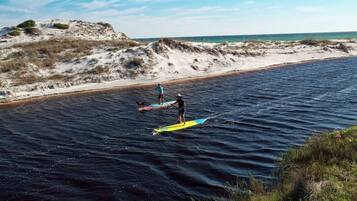 A beach with two paddleboarders on the water, a sandy shore, and dunes in the background.