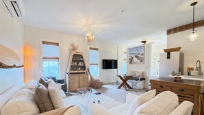A modern living room with white sofas, a wooden dining table, and a kitchen area with a refrigerator and sink.