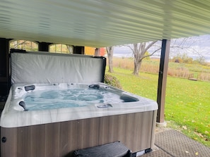A hot tub with a wooden exterior and a transparent cover, situated under a roofed area with a view of a grassy field and trees. - Rent an entire lake-view estate in the heart of wine country!   (Hammondsport)
