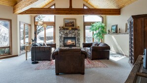 A spacious living room with a stone fireplace, leather sofas, and large windows offering a view of the outdoors. - Panoramic View, Great Outdoor Space, Private Elevator, Monthly Rentals Available (Vail)