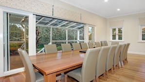 Uma sala de jantar ampla, com uma grande mesa de madeira e várias cadeiras, janelas grandes com vista para árvores, e um design moderno no teto.