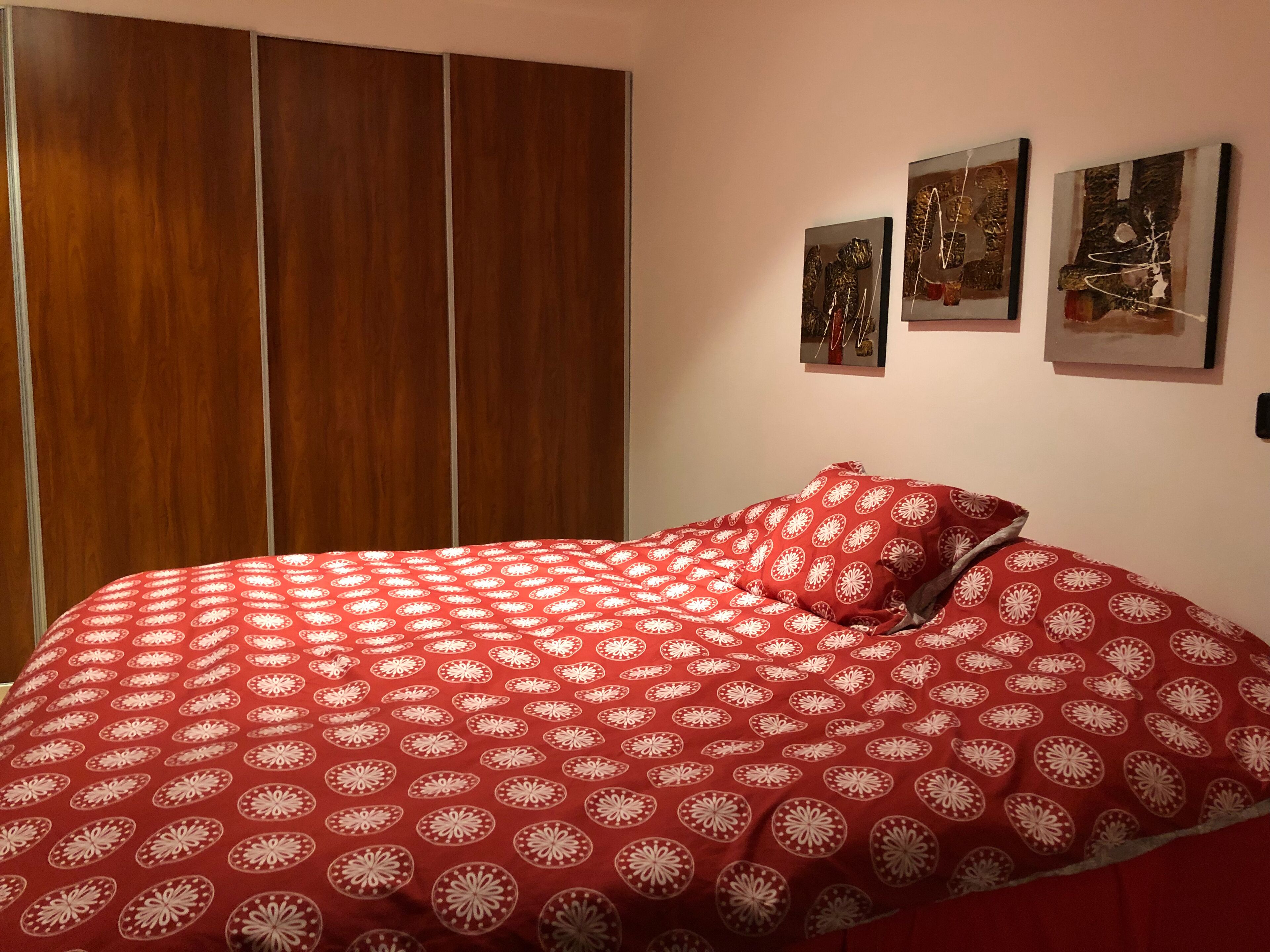 Een slaapkamer met een bed voorzien van een rood beddegoed met patroon, een houten garderobe en drie ingelijste kunstwerken aan de muur.
