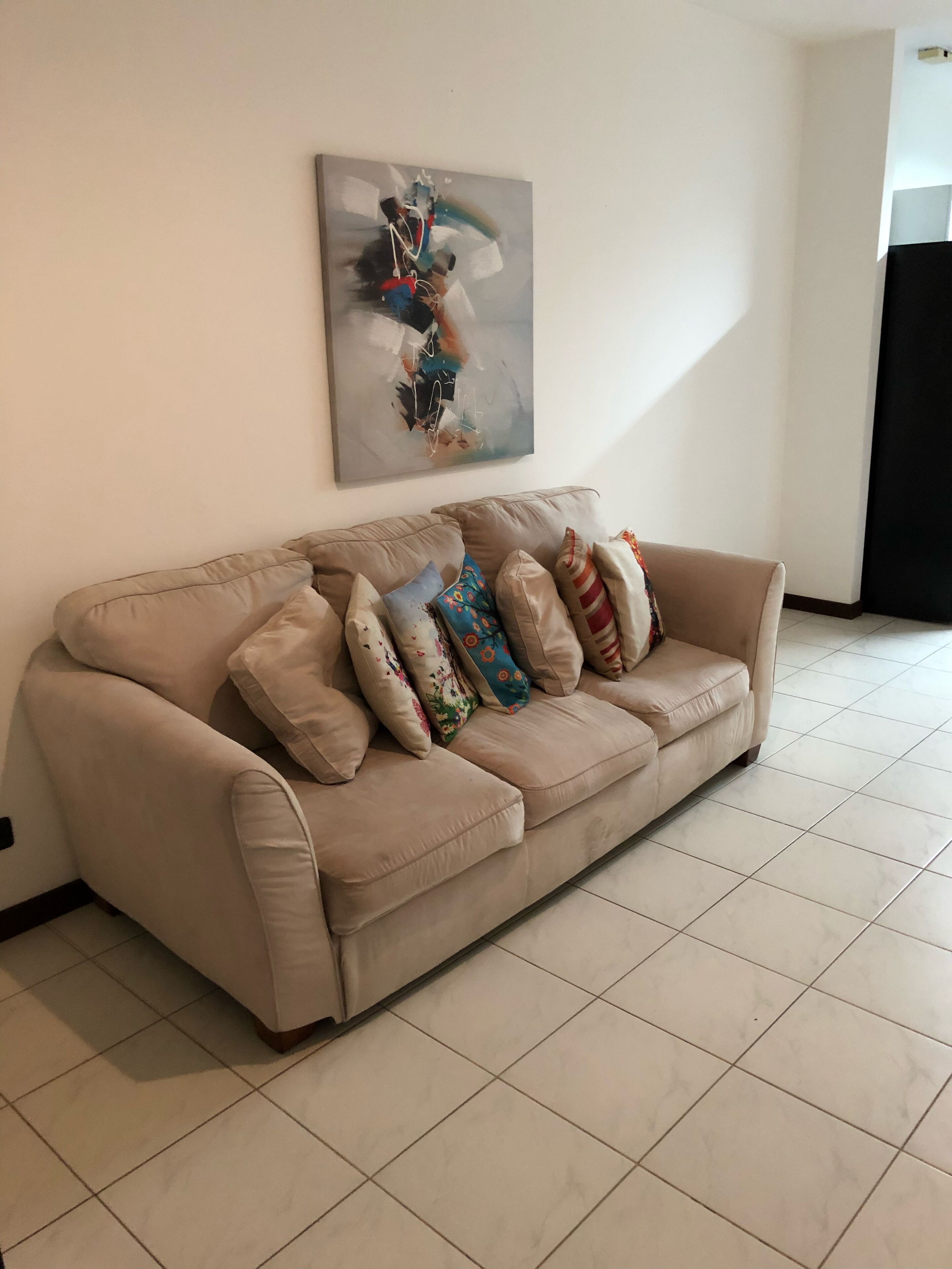 A beige sofa with multiple colorful pillows against a white wall, a tiled floor, and a partial view of a black curtain.