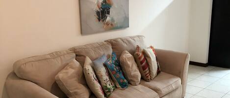 A beige sofa with multiple colorful pillows against a white wall, a tiled floor, and a partial view of a black curtain.